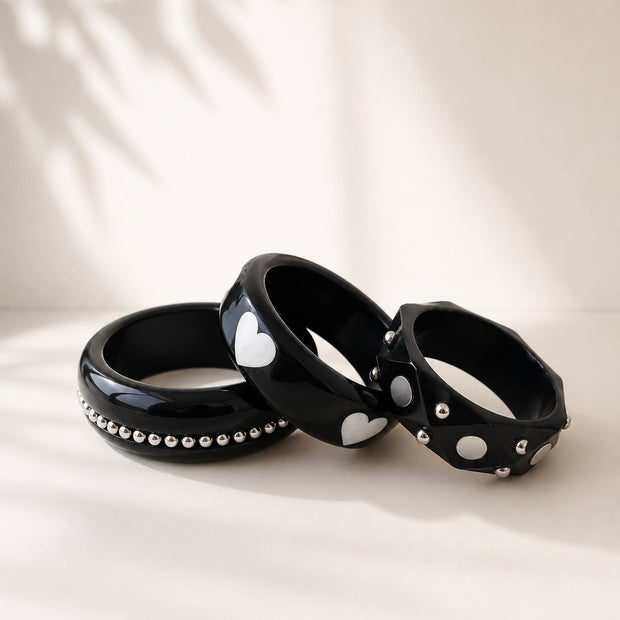 Three black resin bangles with silver studs arranged in a fan on a white surface with leaf shadow