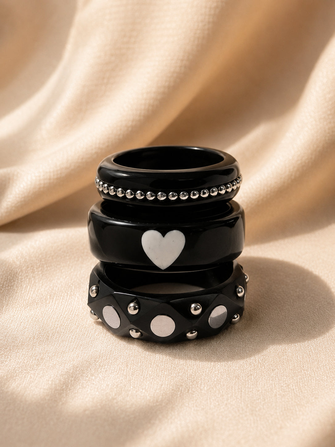 Three black resin bangles stacked vertically showing beaded, heart and studded designs on cream satin