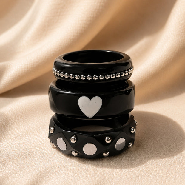 Three black resin bangles stacked vertically showing beaded, heart and studded designs on cream satin