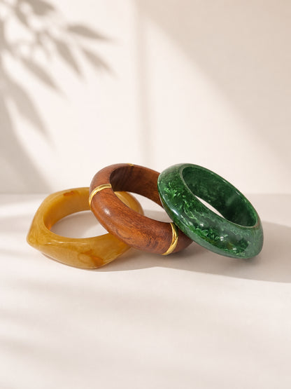 Three stacked bangles in amber resin, polished wood with gold accents, and green marbled resin on white fabric with leaf shadow