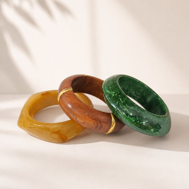 Three stacked bangles in amber resin, polished wood with gold accents, and green marbled resin on white fabric with leaf shadow