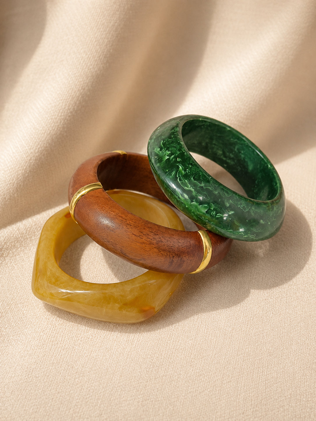 Amber faceted resin bangle, brown wooden bangle with gold band, and green marbled resin bangle grouped on cream fabric from above
