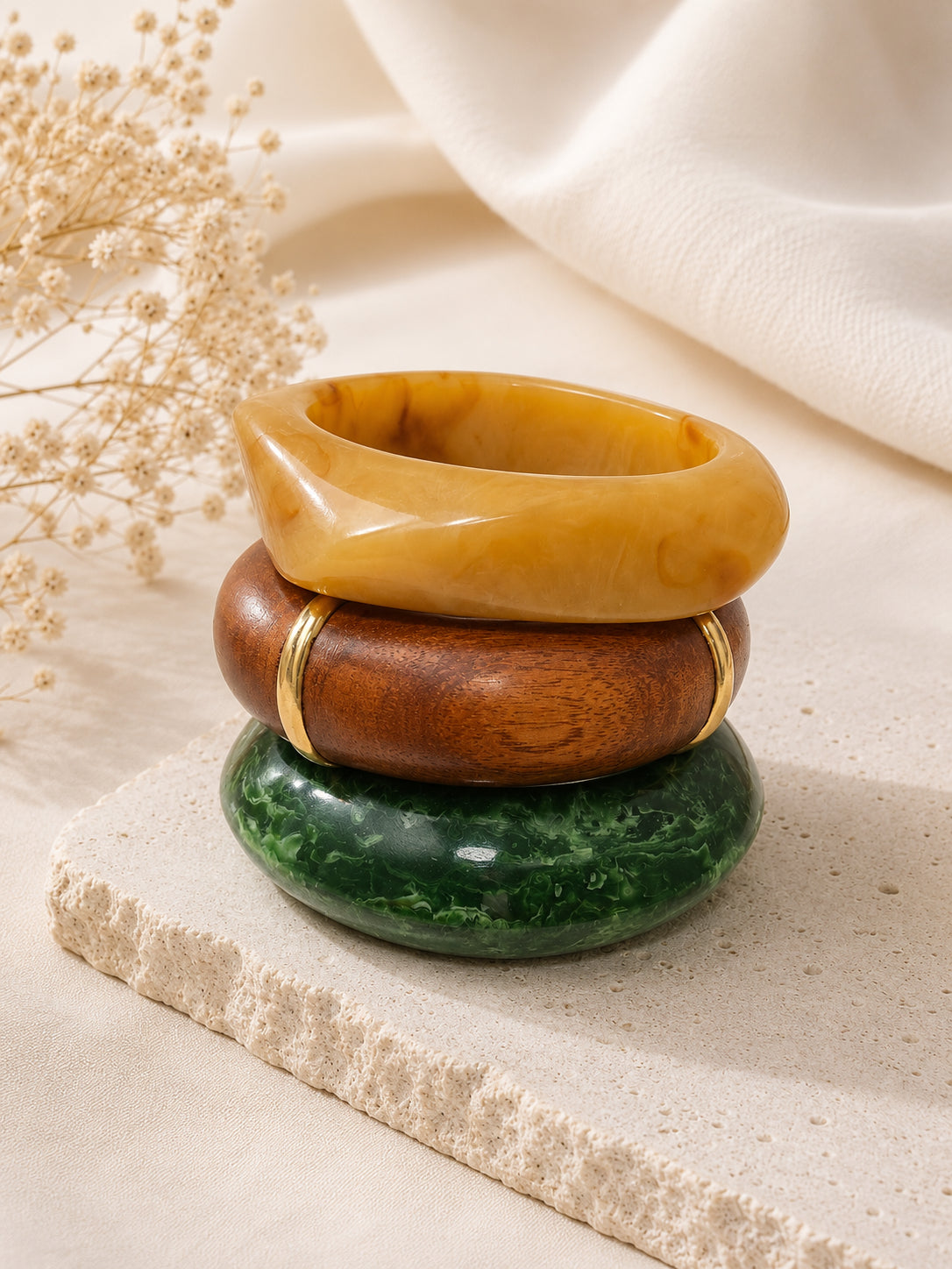 Amber resin, wooden, and green marbled resin bangles stacked on a textured stone surface with dried flowers and white fabric