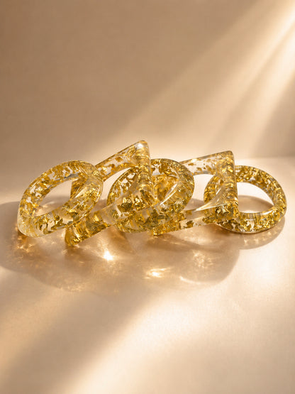 Five clear resin bangles with gold foil flakes scattered loosely on a warm beige surface in soft natural light