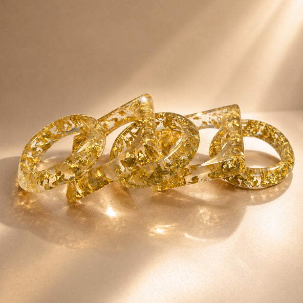 Five clear resin bangles with gold foil flakes scattered loosely on a warm beige surface in soft natural light