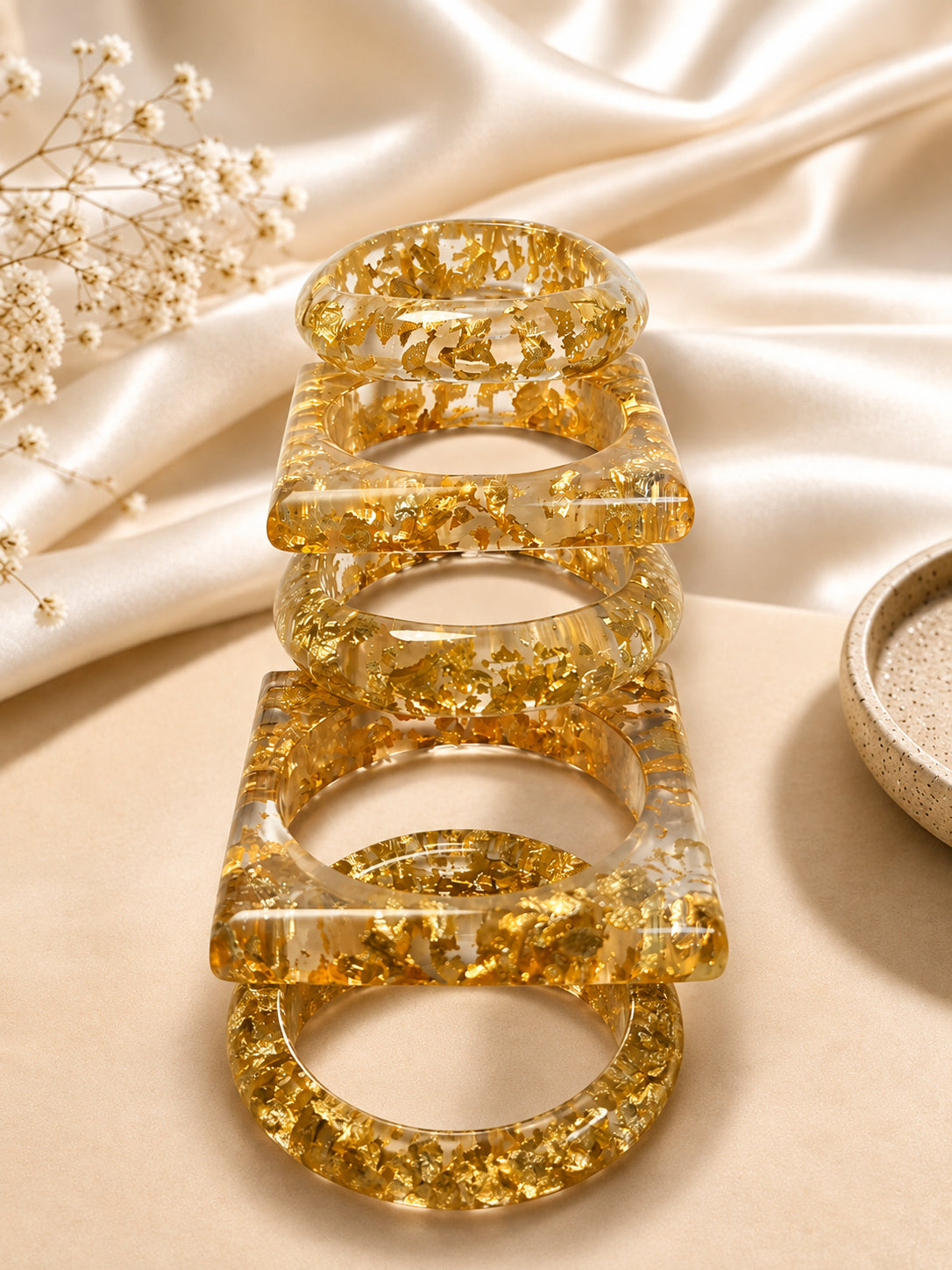 Four gold foil resin bangles stacked upright in a row against a draped white satin cloth with dried flowers
