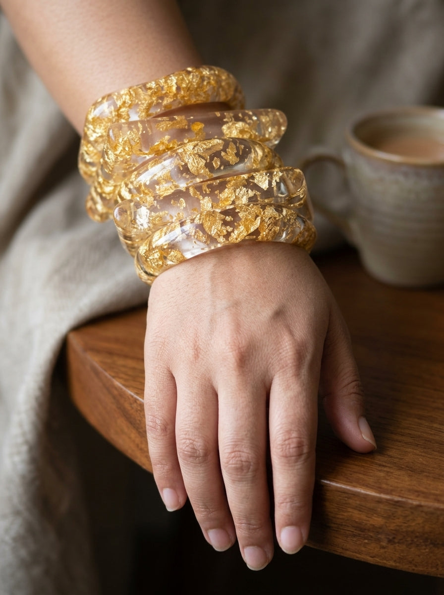 Close-up of a woman's wrist wearing multiple stacked clear resin gold foil bangles resting on a wooden table