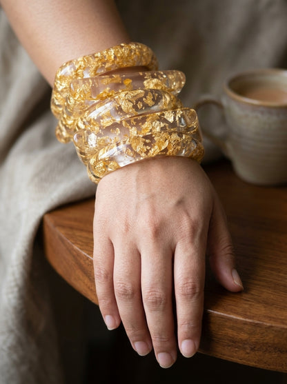 Close-up of a woman's wrist wearing multiple stacked clear resin gold foil bangles resting on a wooden table