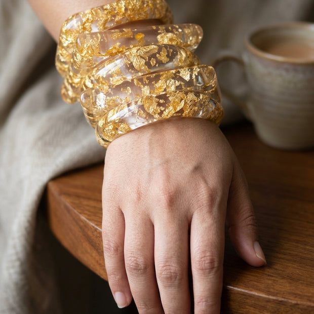 Close-up of a woman's wrist wearing multiple stacked clear resin gold foil bangles resting on a wooden table