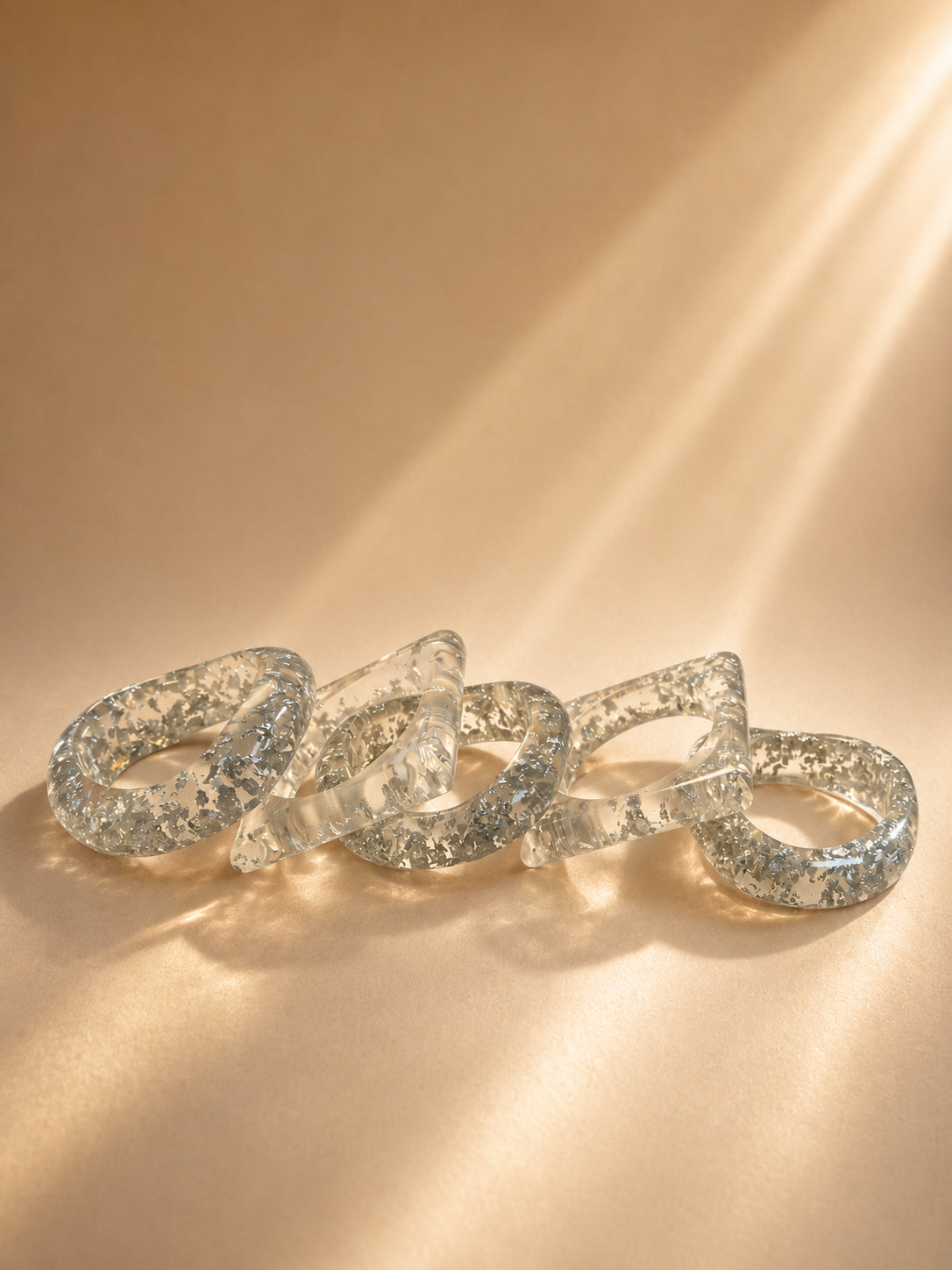Five clear resin bangles with silver foil flakes laid flat on a warm beige surface in soft light