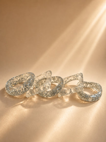 Five clear resin bangles with silver foil flakes laid flat on a warm beige surface in soft light