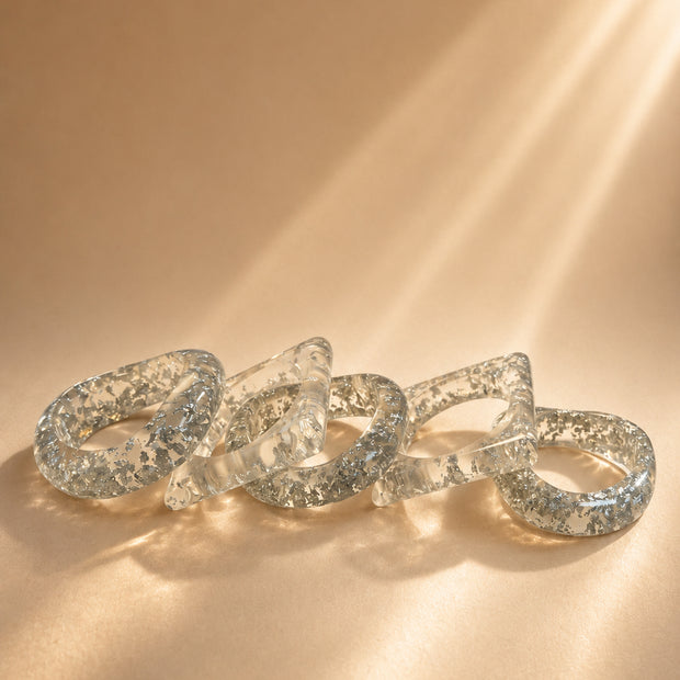 Five clear resin bangles with silver foil flakes laid flat on a warm beige surface in soft light