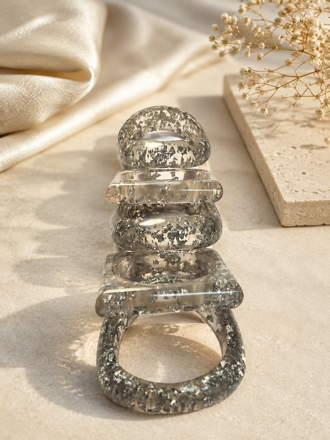 Five silver foil resin bangles stacked upright on a stone surface beside dried flowers and silk fabric