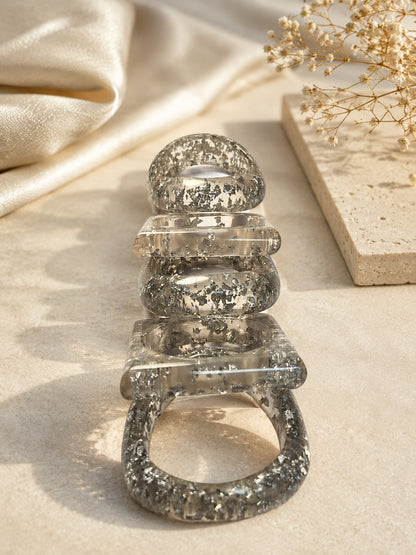 Five silver foil resin bangles stacked upright on a stone surface beside dried flowers and silk fabric