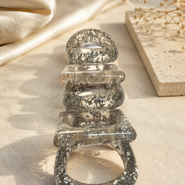 Five silver foil resin bangles stacked upright on a stone surface beside dried flowers and silk fabric