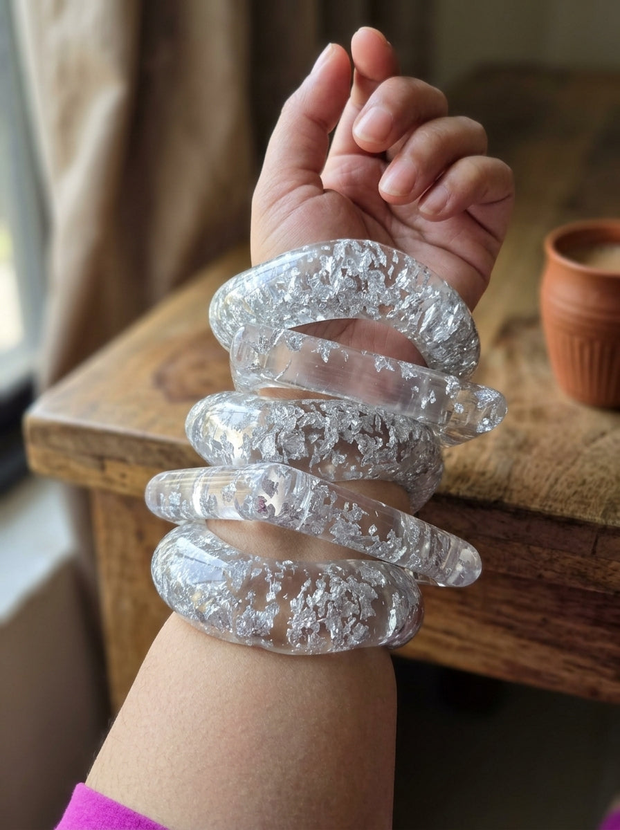 Multiple clear silver foil resin bangles stacked on a woman's wrist against a wooden table background