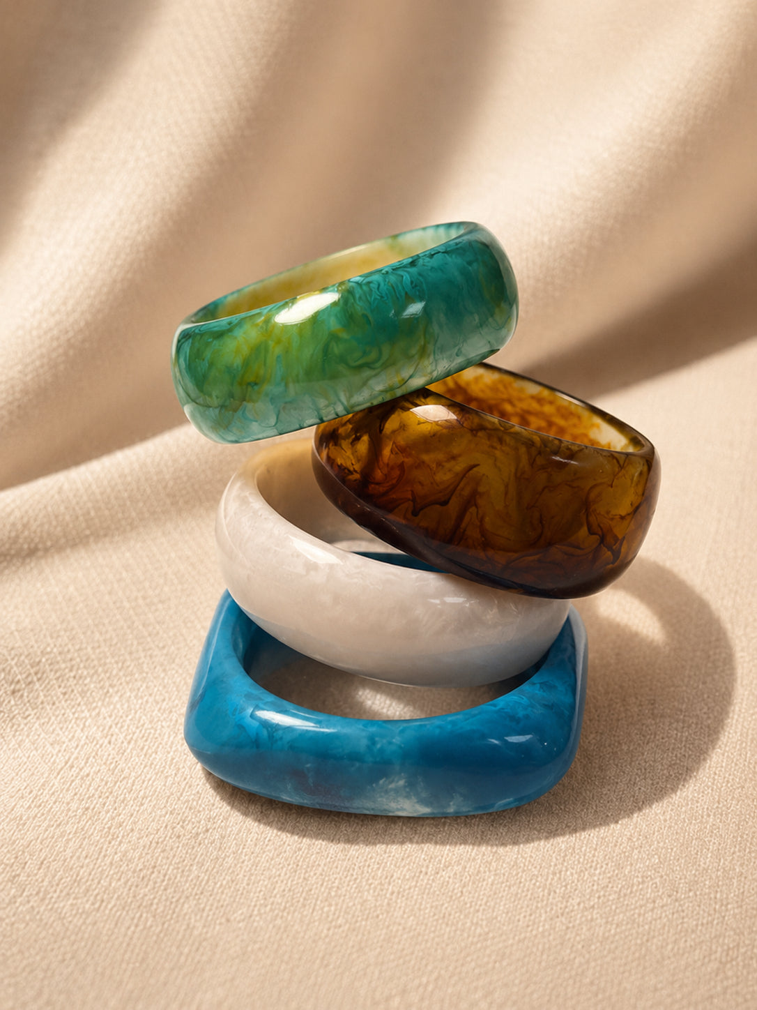Four resin marble bangles stacked at an angle on cream fabric showing teal, amber, white and blue finishes