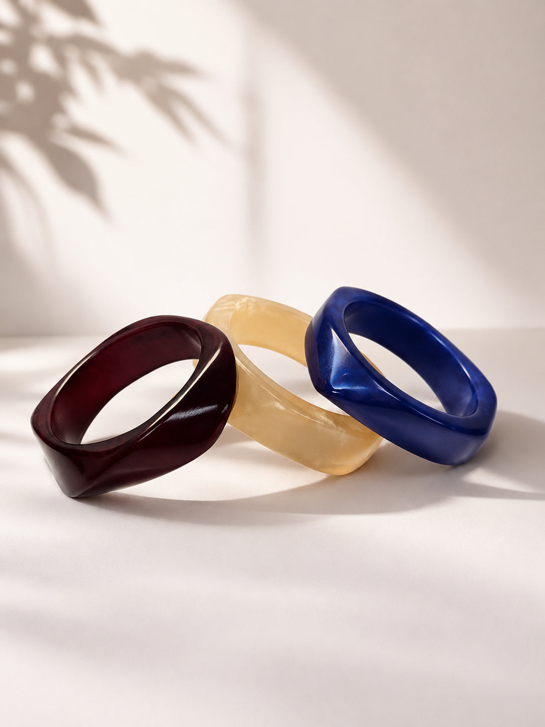 Three resin bangles in burgundy, ivory and cobalt blue arranged in a fan on a white surface