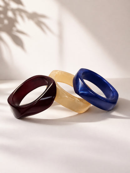 Three resin bangles in burgundy, ivory and cobalt blue arranged in a fan on a white surface