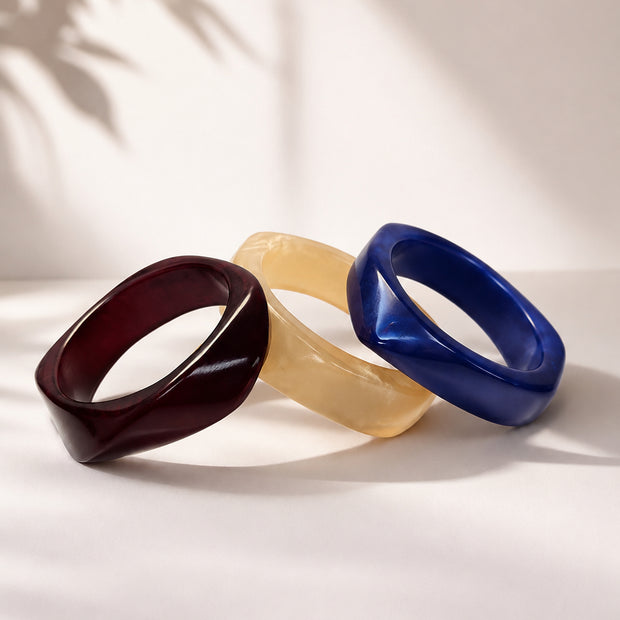 Three resin bangles in burgundy, ivory and cobalt blue arranged in a fan on a white surface