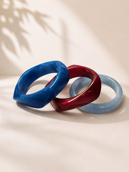 Three marble resin bangles in cobalt blue, deep burgundy, and powder blue arranged side by side on a cream surface