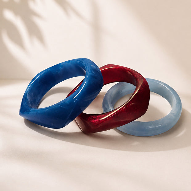 Three marble resin bangles in cobalt blue, deep burgundy, and powder blue arranged side by side on a cream surface