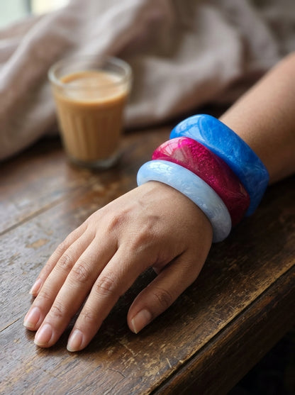Woman wearing all three marble resin bangles stacked on her wrist resting on a wooden table beside a glass of chai