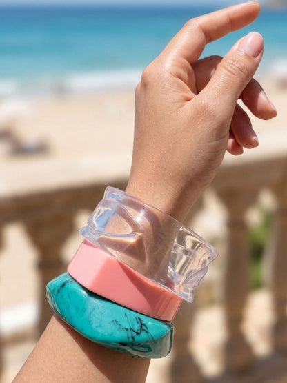 Three stacked resin bangles on a raised wrist against a blurred beach and stone balustrade background