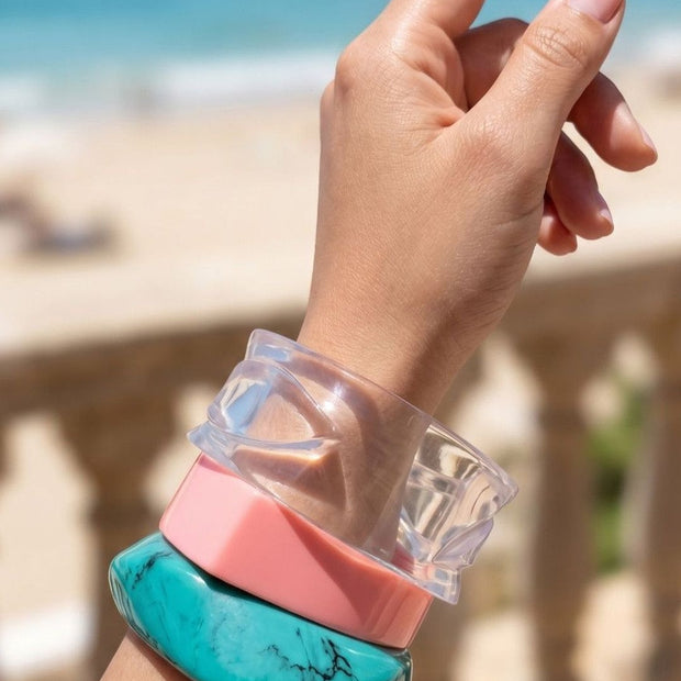 Three stacked resin bangles on a raised wrist against a blurred beach and stone balustrade background