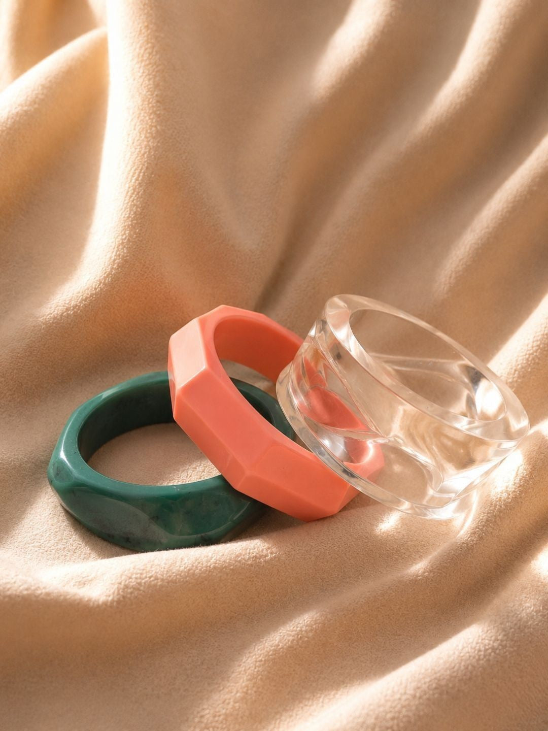 Three resin bangles laid flat on draped golden fabric showing clear, coral and green styles