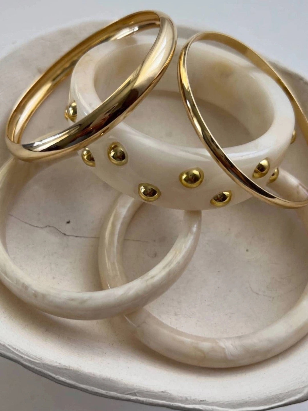 Three bangles from the set arranged on a white ceramic dish — studded cuff, plain resin and gold metal bangle
