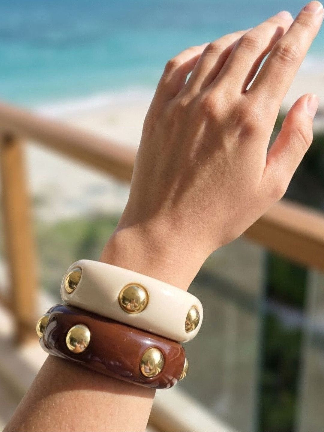 Both gold-studded resin bangles worn stacked on a wrist against a bright ocean backdrop
