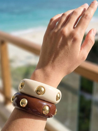 Both gold-studded resin bangles worn stacked on a wrist against a bright ocean backdrop