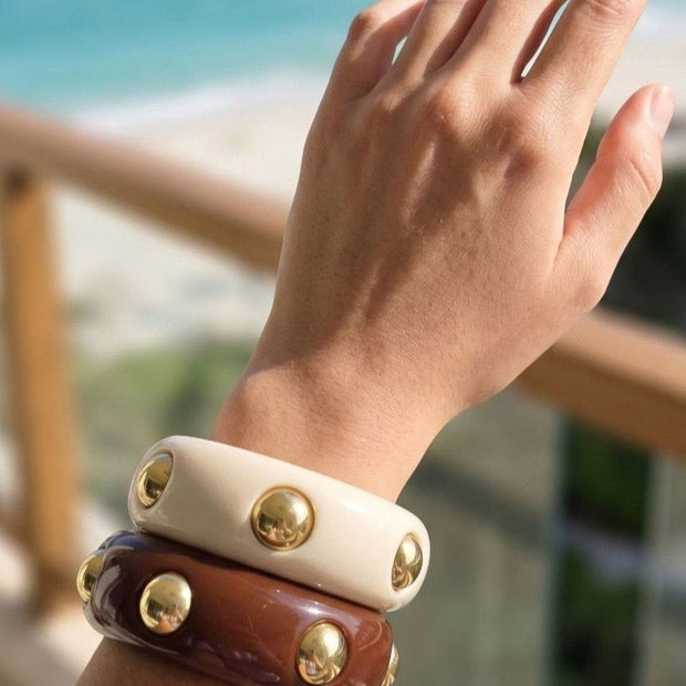 Both gold-studded resin bangles worn stacked on a wrist against a bright ocean backdrop
