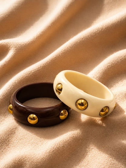 Ivory and dark brown gold dome-stud resin bangles laid side by side on draped sand-toned fabric