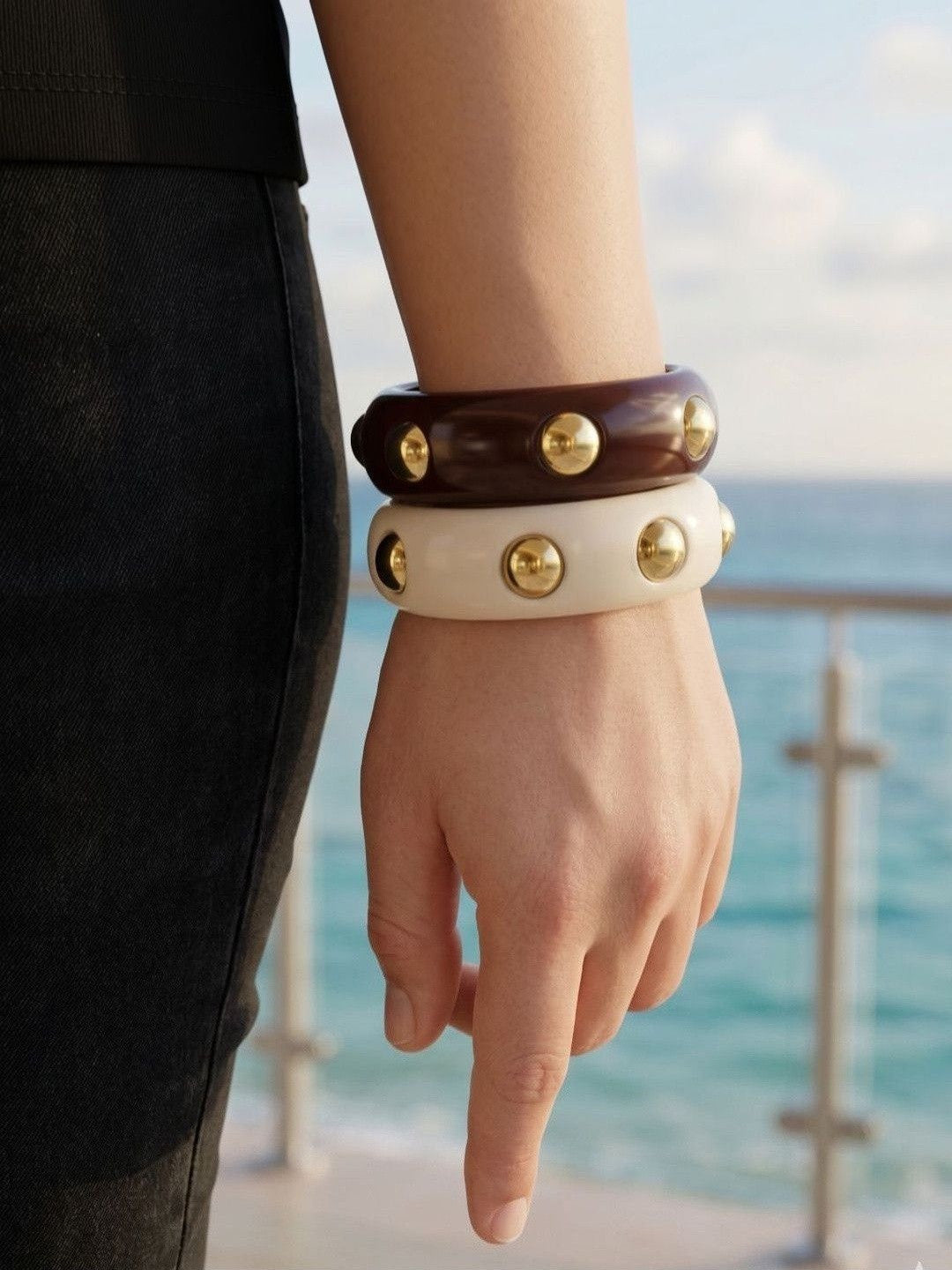 Stacked ivory and brown gold-studded resin bangles on a wrist with a black outfit and sea in background
