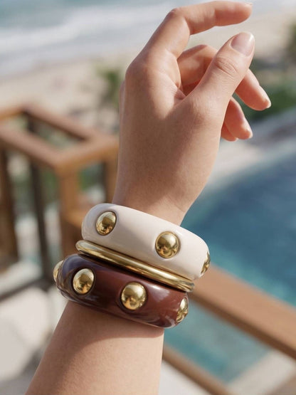 Three resin bangles stacked on a wrist — cream and burgundy with gold dome studs, shot against a beach backdrop
