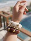 Three resin bangles stacked on a wrist — cream and burgundy with gold dome studs, shot against a beach backdrop
