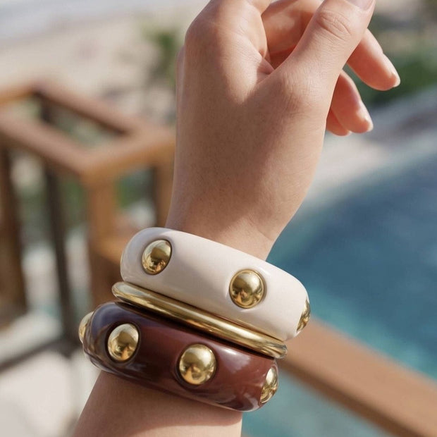 Three resin bangles stacked on a wrist — cream and burgundy with gold dome studs, shot against a beach backdrop
