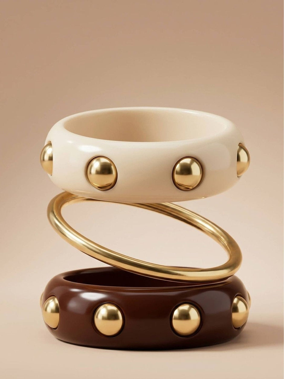 Cream resin bangle with gold studs stacked above a gold-tone slim bangle and a burgundy stud bangle on a neutral background