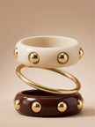 Cream resin bangle with gold studs stacked above a gold-tone slim bangle and a burgundy stud bangle on a neutral background