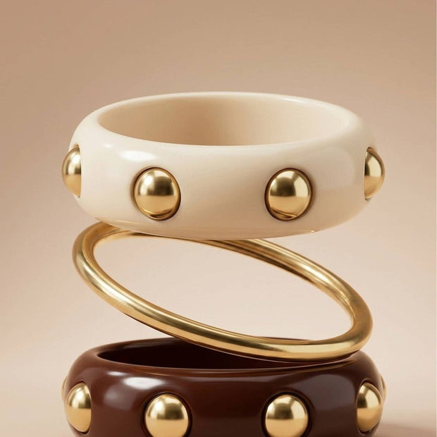 Cream resin bangle with gold studs stacked above a gold-tone slim bangle and a burgundy stud bangle on a neutral background