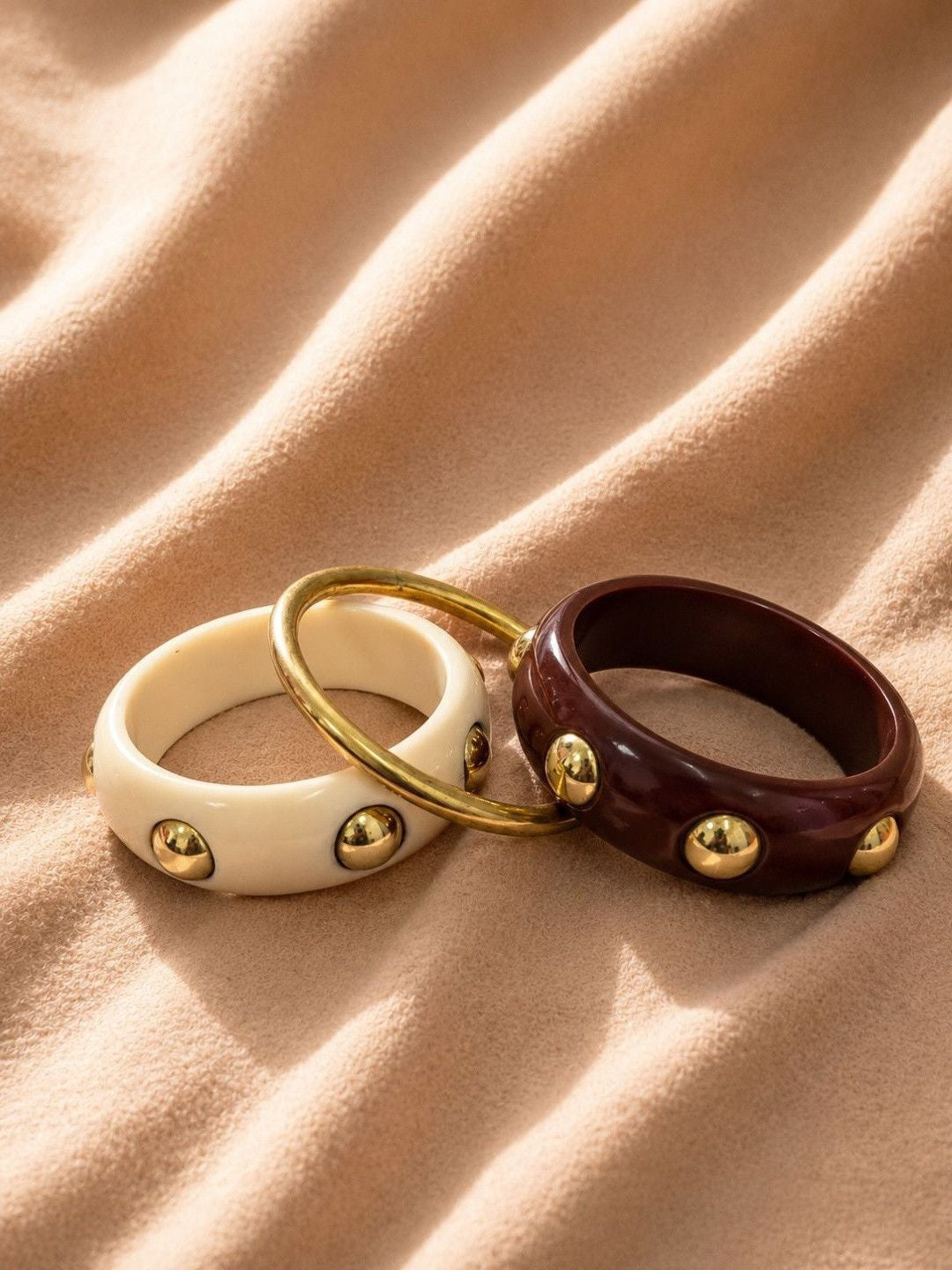 Cream, gold-tone and burgundy bangles laid flat on a warm sandy fabric surface showing stud detail
