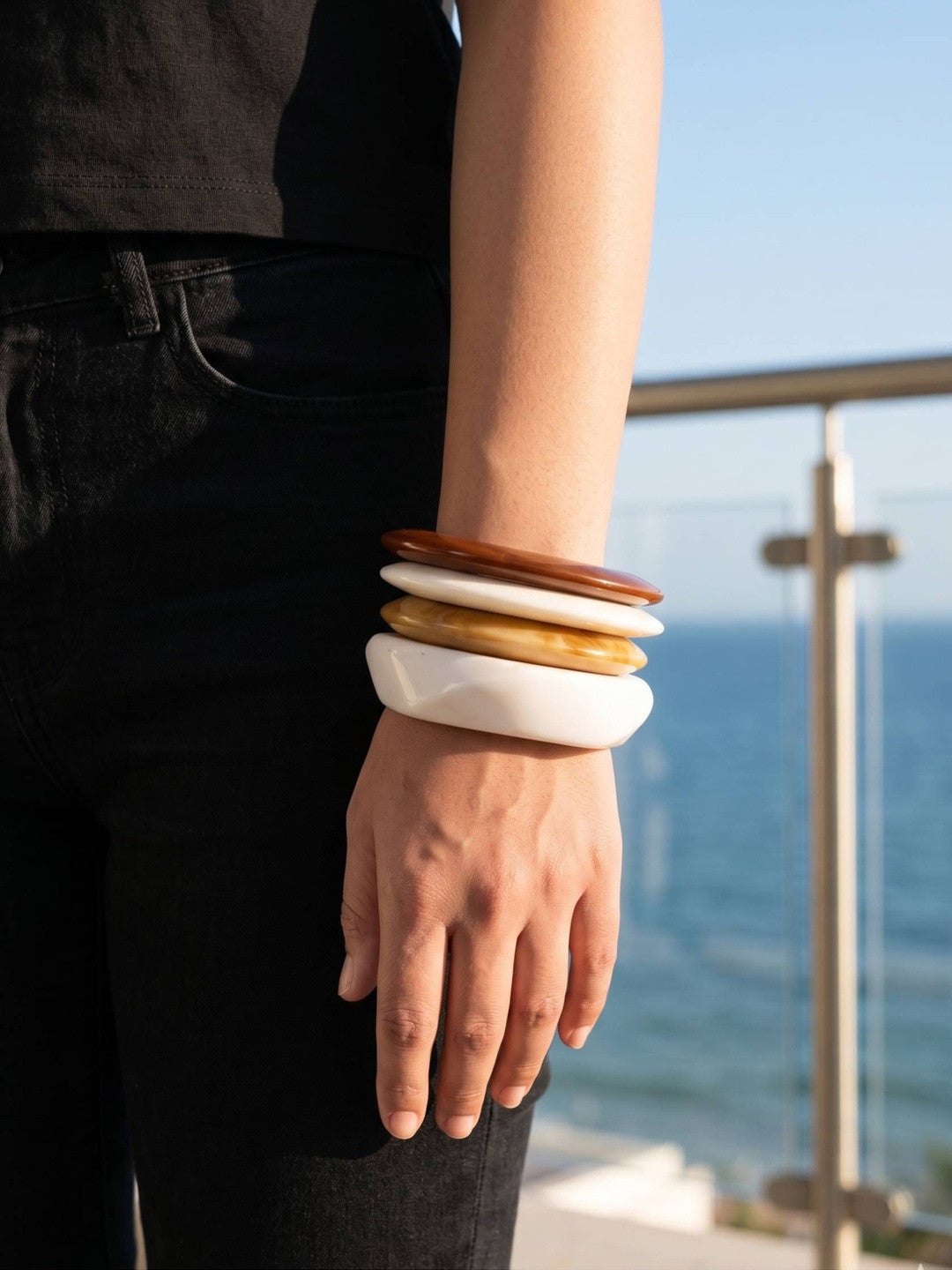 Four resin bangles stacked on a wrist, terracotta on top grading down to white, worn with black outfit against an ocean backdrop