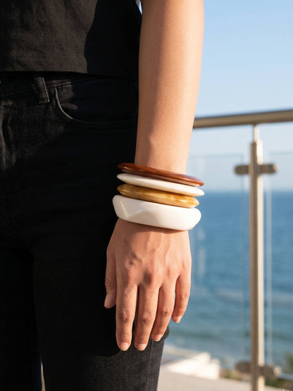 Four resin bangles stacked on a wrist, terracotta on top grading down to white, worn with black outfit against an ocean backdrop