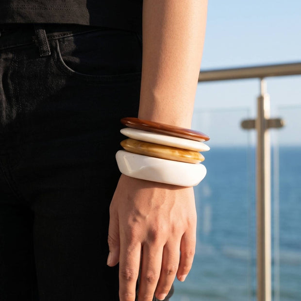 Four resin bangles stacked on a wrist, terracotta on top grading down to white, worn with black outfit against an ocean backdrop