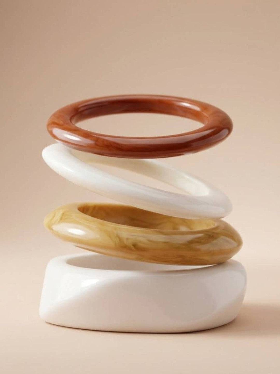 Four glossy resin bangles arranged in a floating sculptural stack showing terracotta, cream, amber and white profiles on a beige background
