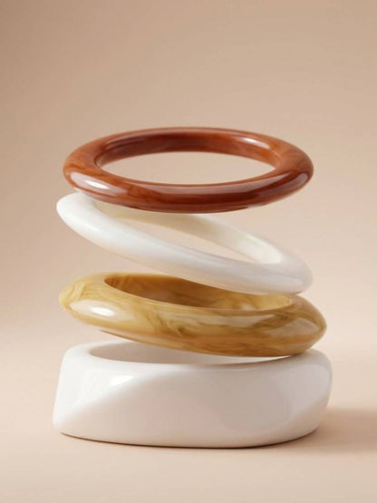 Four glossy resin bangles arranged in a floating sculptural stack showing terracotta, cream, amber and white profiles on a beige background