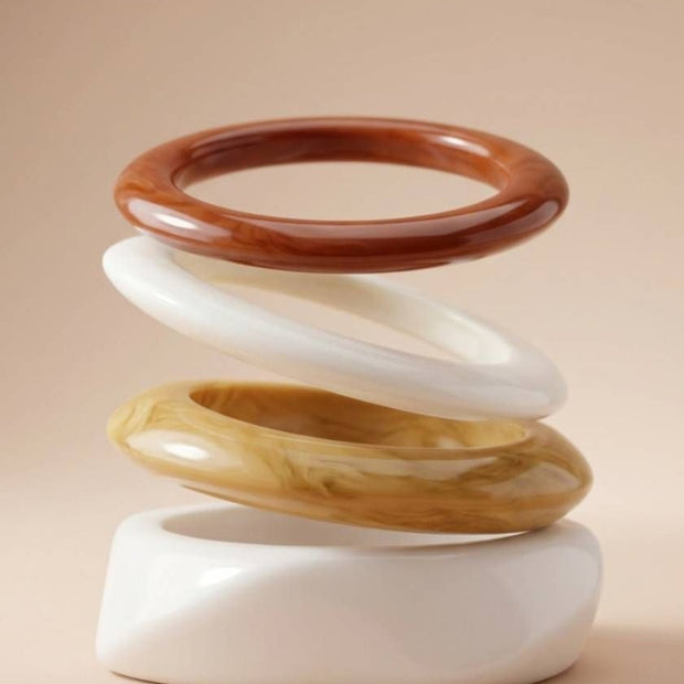 Four glossy resin bangles arranged in a floating sculptural stack showing terracotta, cream, amber and white profiles on a beige background
