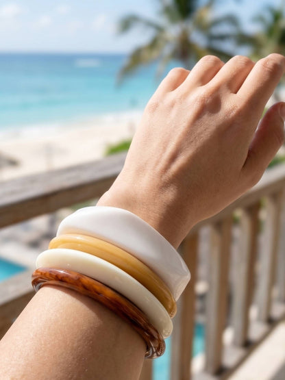 All four resin bangles worn together on a wrist held up near a tropical beachside balcony railing in bright sunlight